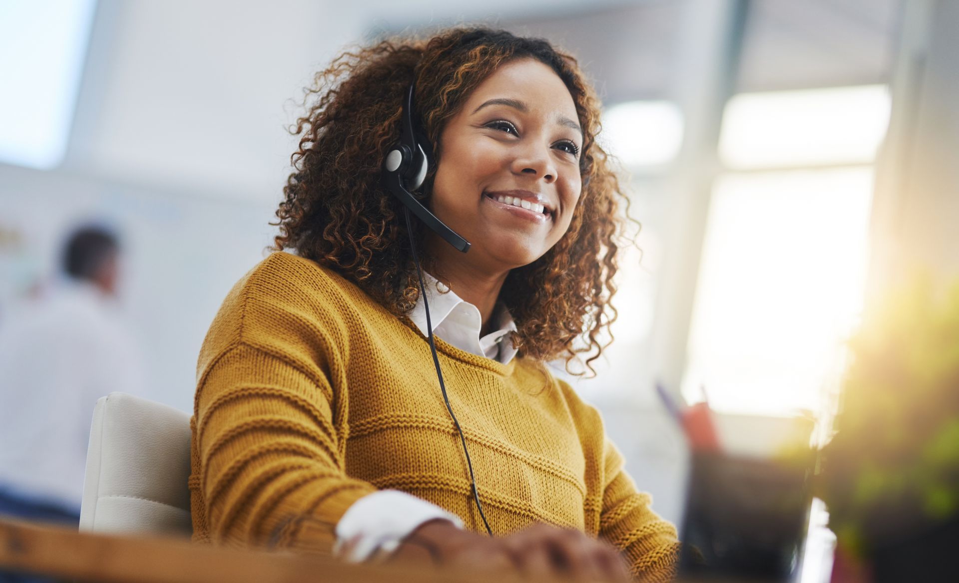 Woman with curly hair wearing a headset, smiling, sitting at a desk with a warm, sunny background.