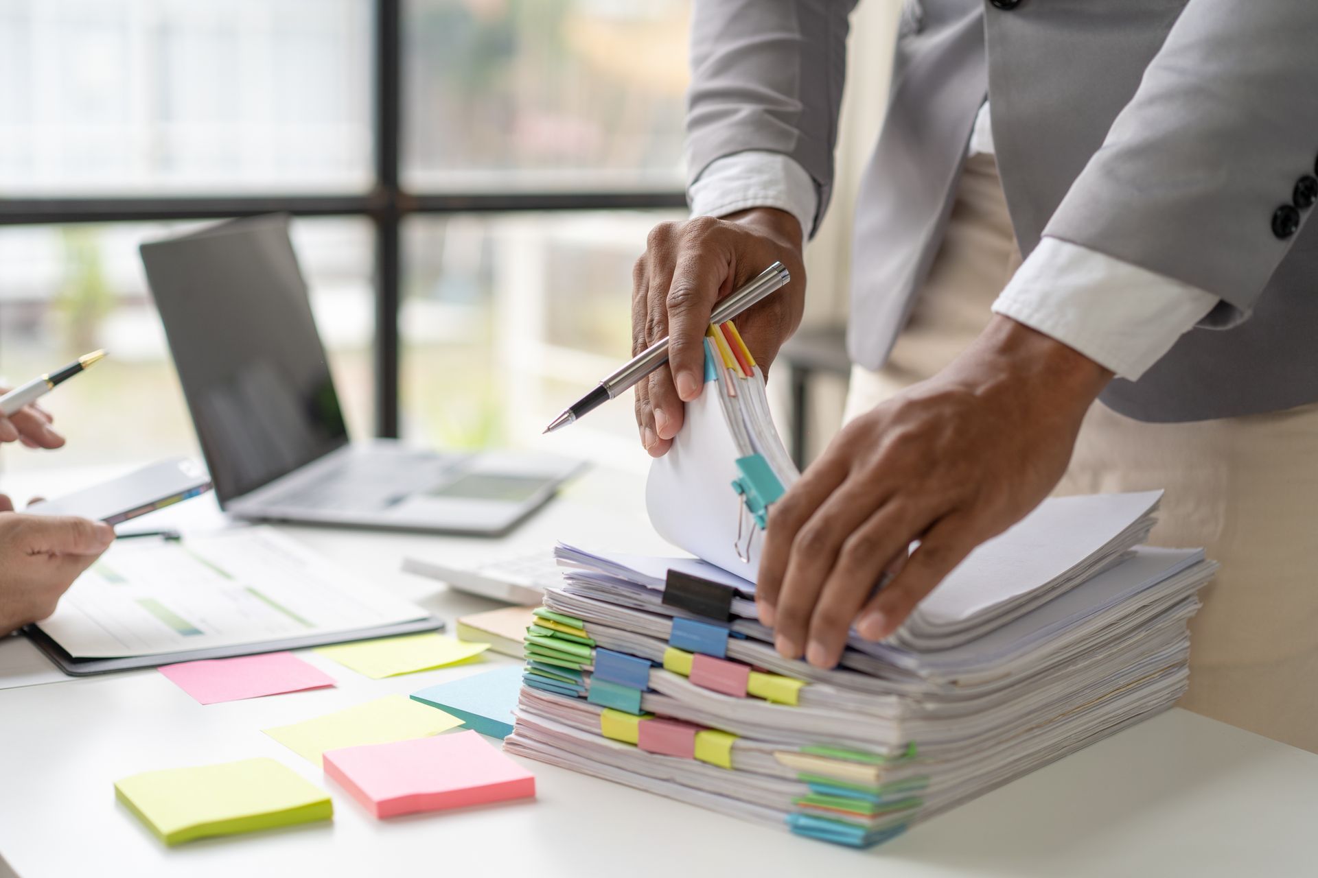 Person sorting through documents, stacks of papers on desk with sticky notes and laptop.