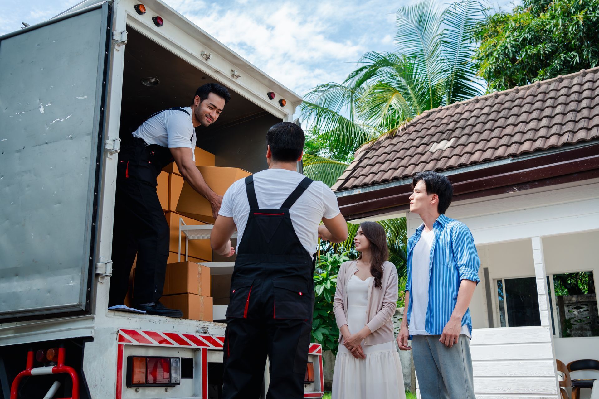 Moving truck being loaded with boxes; couple watches movers outside a house.