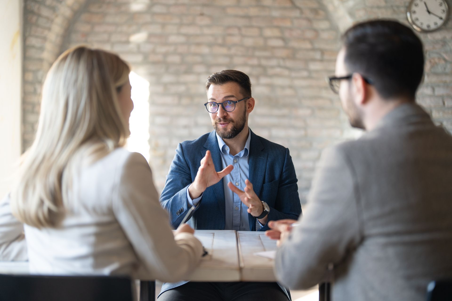 Three people at a table; one man gestures as he speaks, facing two others. Brick wall backdrop.