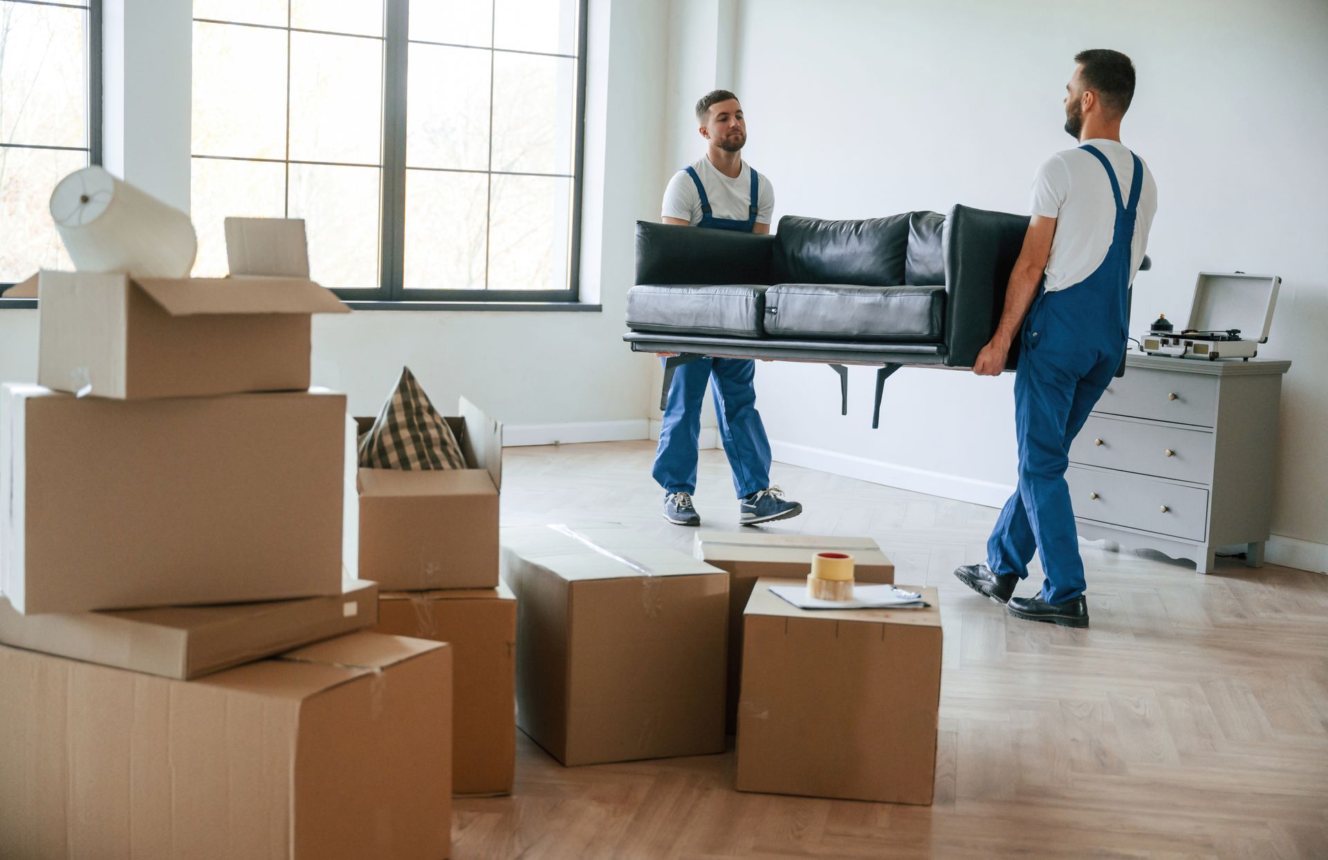 Two movers in blue coveralls carrying a black sofa into a mostly empty room with boxes.