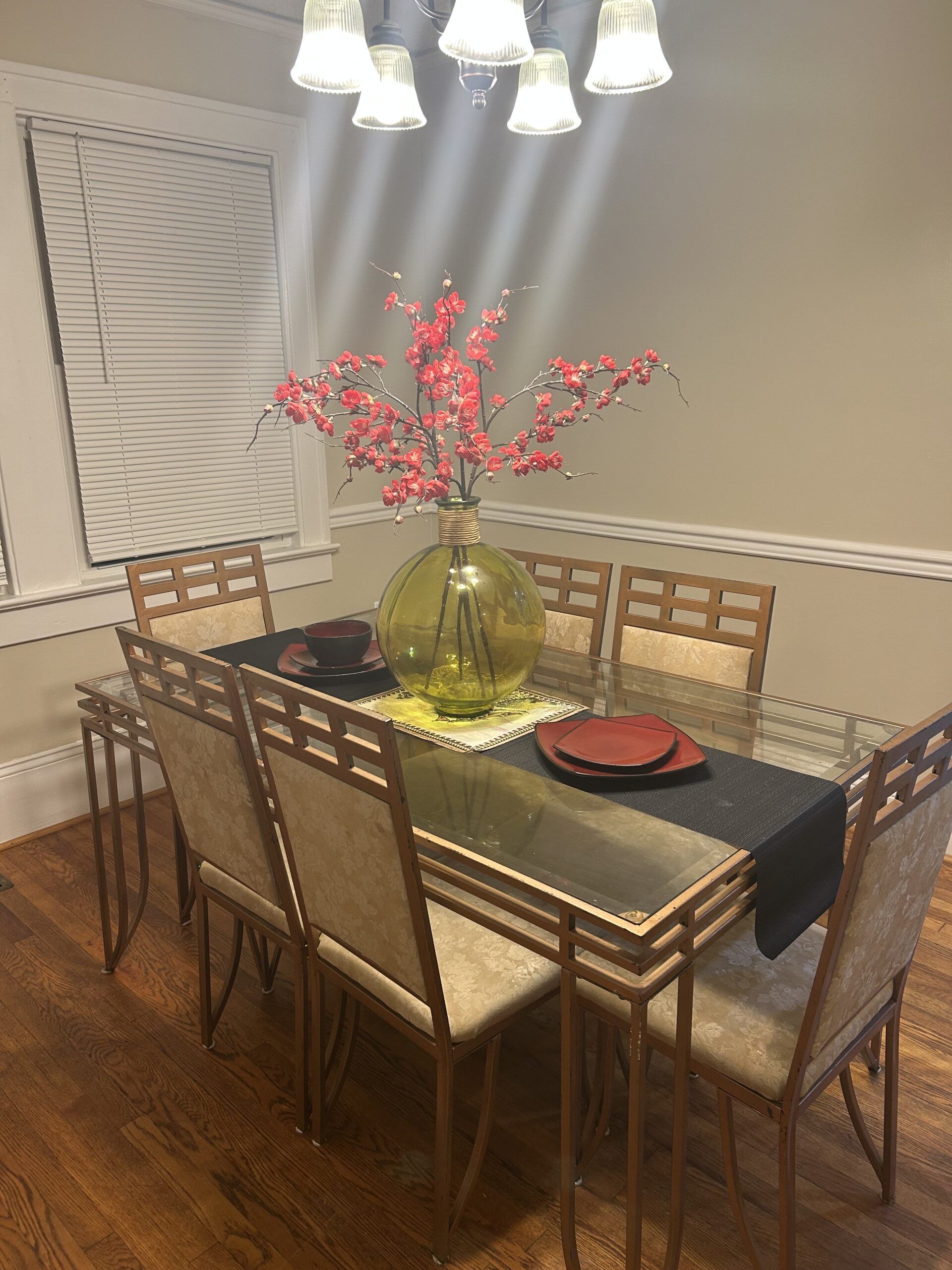 Dining room with a table set with red and black accents, a large green vase with pink branches, and beige chairs.
