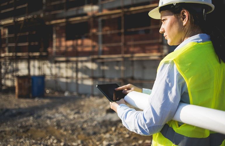 Construction worker in a hard hat and safety vest using a tablet at a building site.