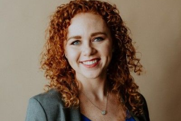 Headshot of a smiling person with curly auburn hair, wearing a grey blazer, against a neutral background.