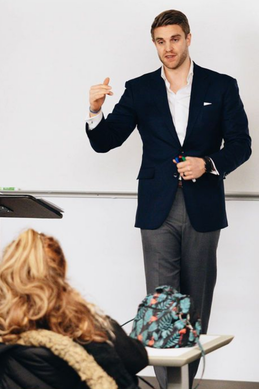 A person in a navy blazer stands by a whiteboard, gesturing while speaking to a group in a classroom.