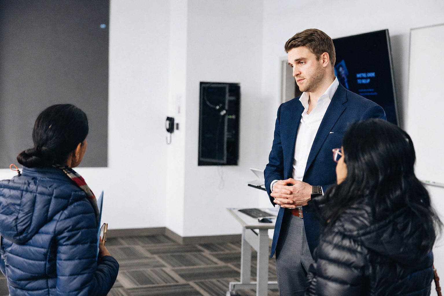 Three people talking in a modern office lobby, with two in dark jackets facing a man in a navy blazer.