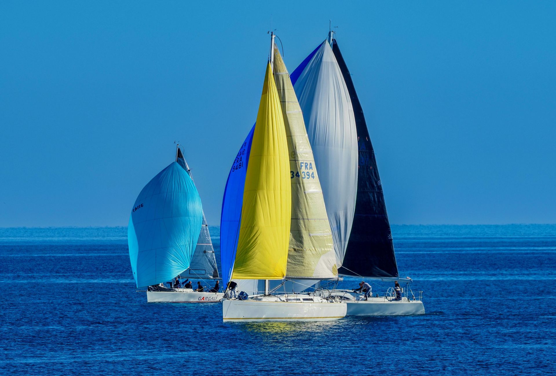 Three sailboats with vibrant blue, yellow, and white spinnaker sails racing on calm blue water.