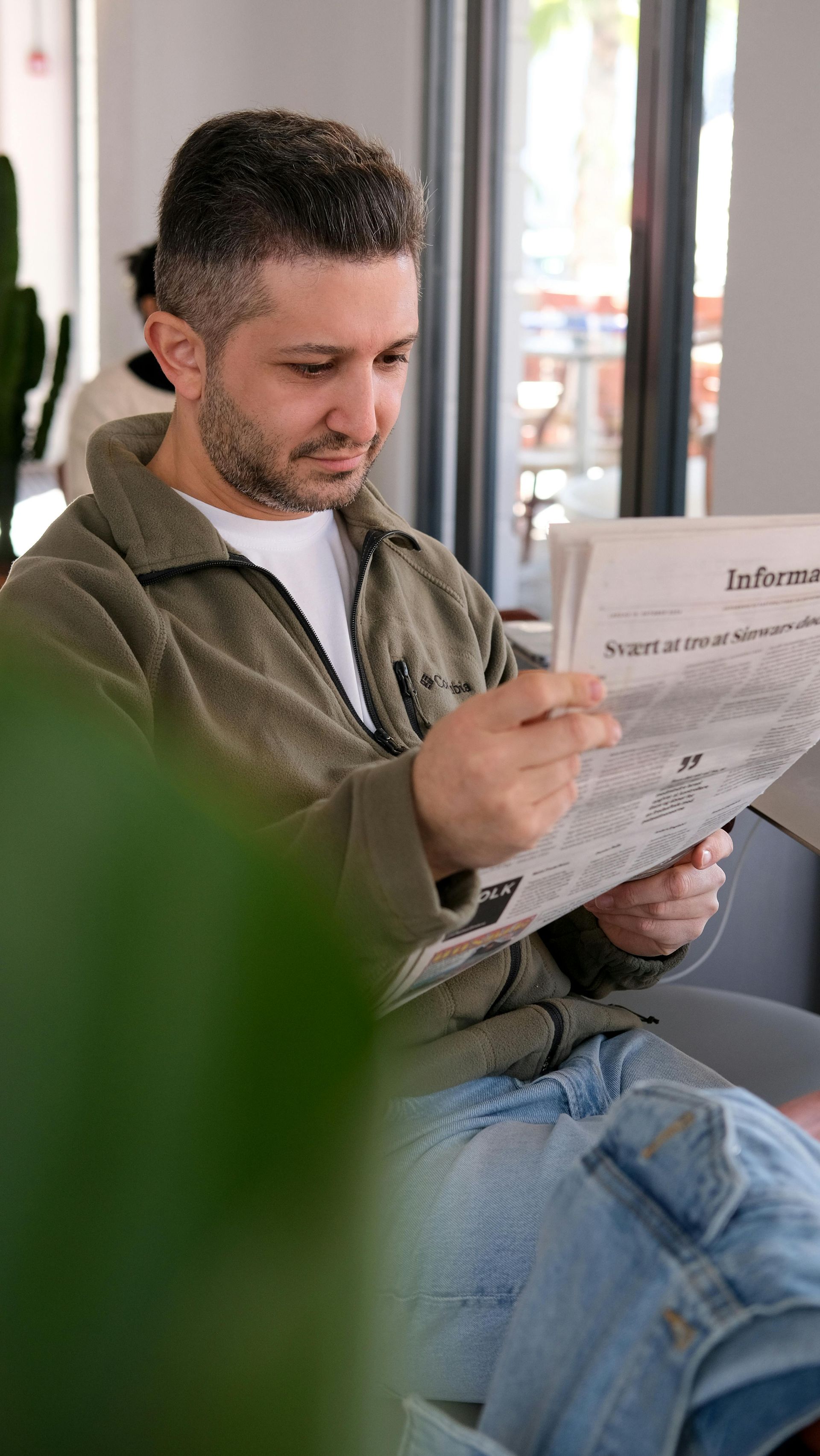 A person with short hair, wearing a green fleece zip-up jacket, sits indoors reading a newspaper.