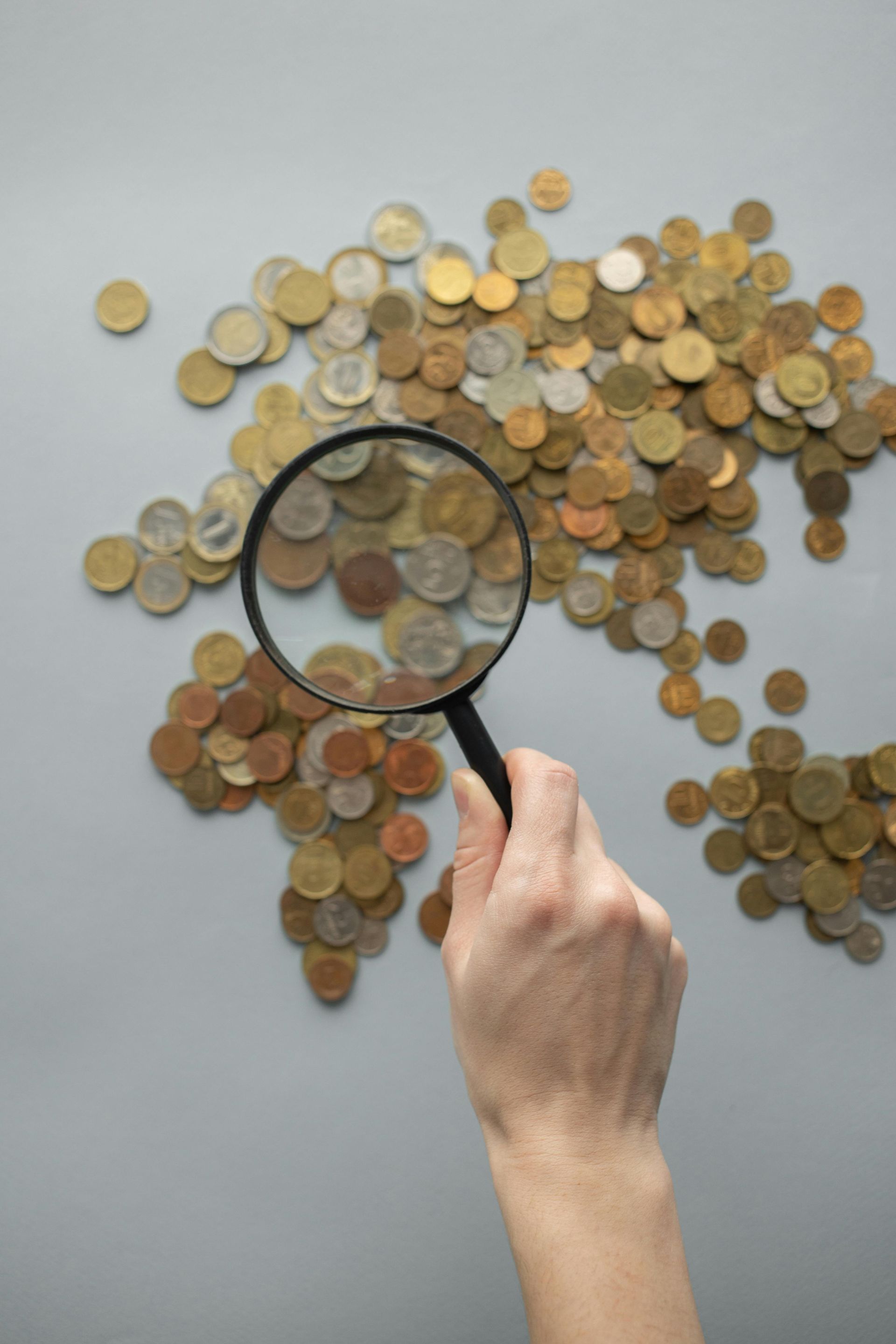A hand holds a magnifying glass over coins arranged to resemble a map of the world against a light gray background.