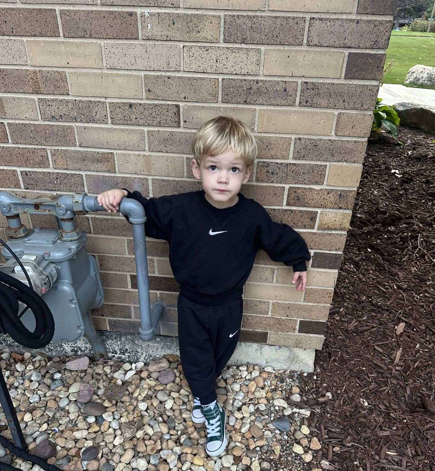 Boy in black Nike sweatsuit leaning on a pipe against a brick wall, pebbles on the ground.