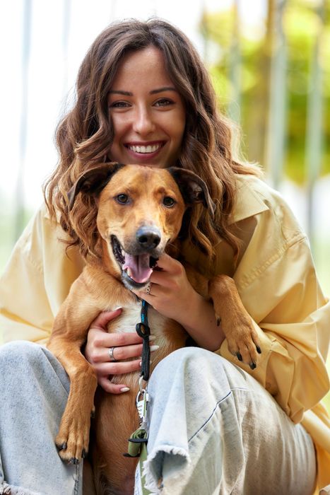 Woman sitting with a brown dog, both smiling. The dog is panting, and the woman is wearing a yellow shirt.
