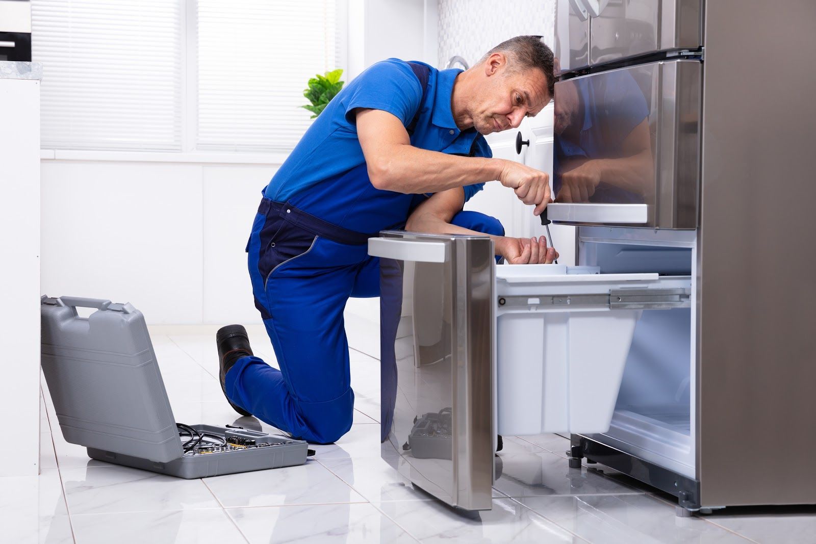 Technician in blue coveralls repairing a stainless-steel appliance with tools in a bright kitchen-like room