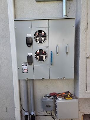 A dual-meter electrical panel mounted on a beige wall with a smaller junction box and tools resting below it.
