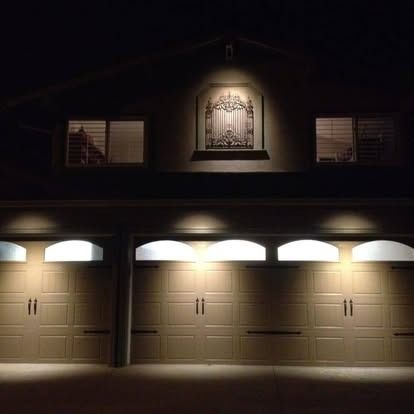 A residential house exterior at night, featuring three garage doors and two illuminated upper-level windows.