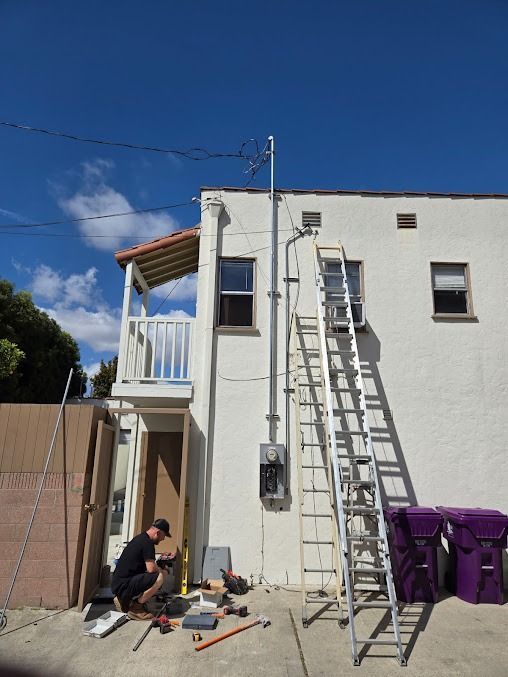 A worker kneels on a concrete driveway while installing an electrical service mast and meter box on an exterior building.