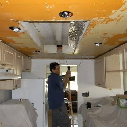 A person stands on a ladder using a power tool to work on an opening in a kitchen ceiling with orange paint and lighting.