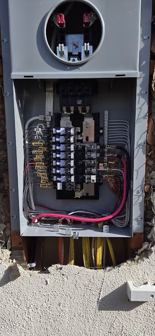 An open electrical meter base and service panel installed on an exterior wall with various conduits and wiring visible.
