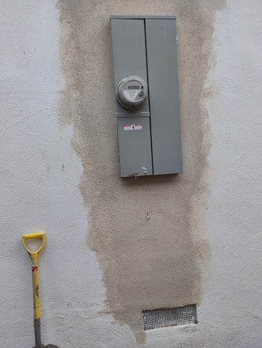An electrical meter panel mounted on a light-colored stucco wall with a visible damp stain and a yellow shovel nearby.