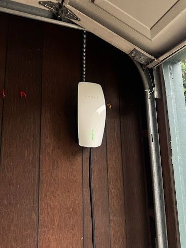 A white Tesla wall charger mounted on a wooden garage wall with a green status light illuminated.