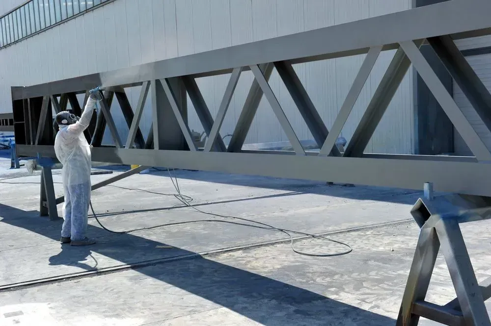 Person in protective suit spray-painting a large gray metal truss structure outdoors.