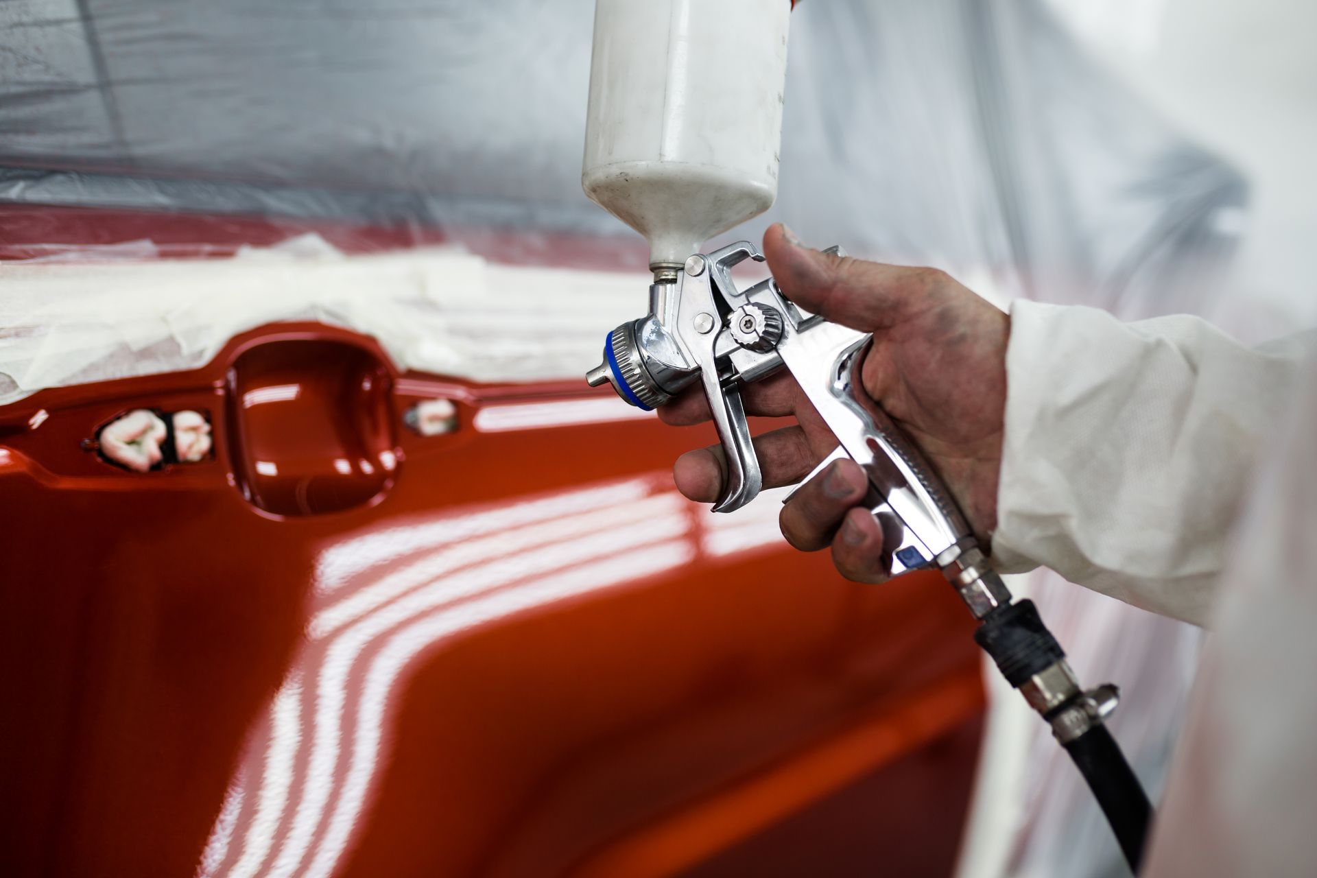 Person in protective suit using a paint sprayer to apply red paint to a car door.