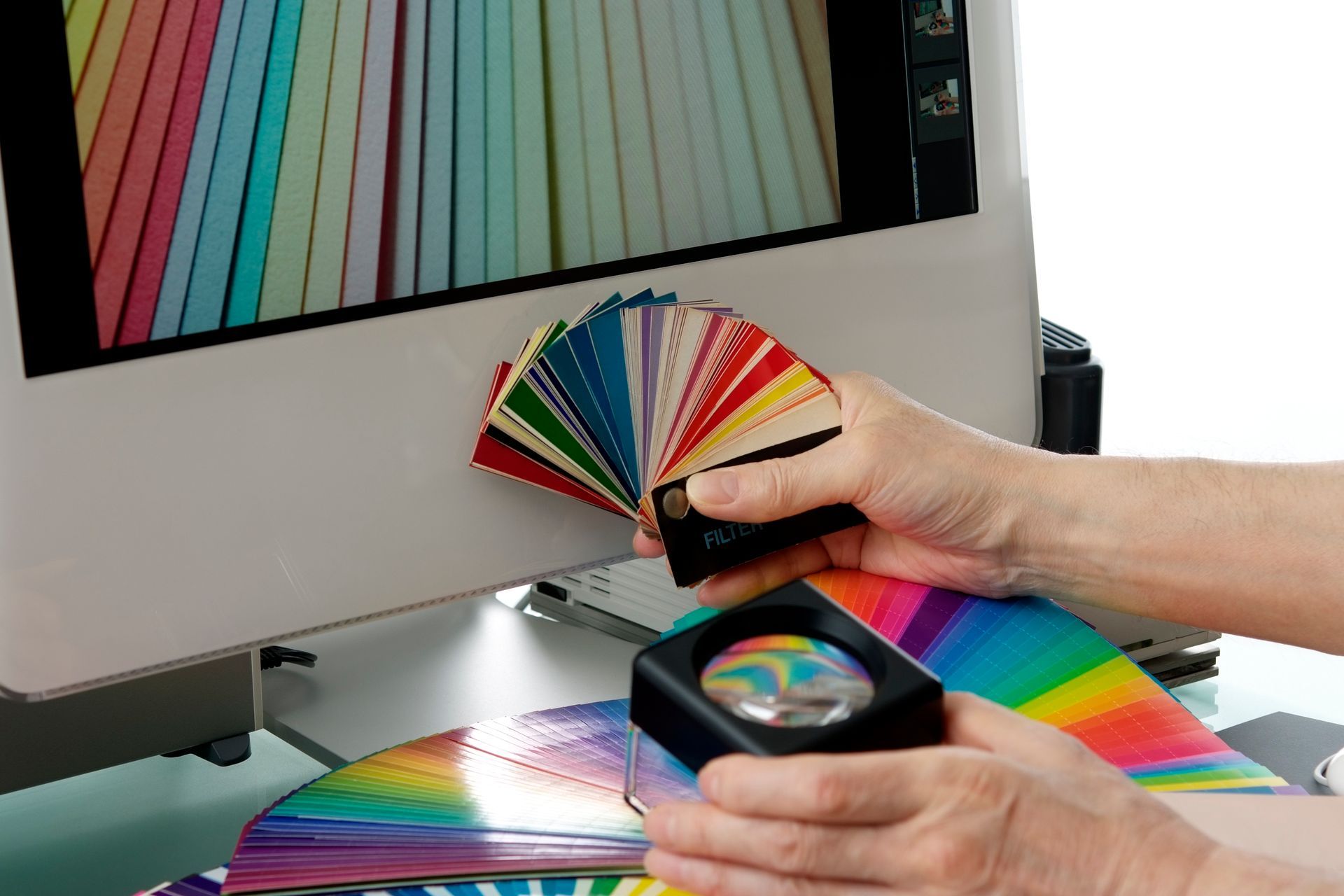 Hands holding a color palette fan and a magnifying glass, comparing colors with a monitor displaying a color range.