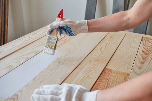 Person painting white paint on a wooden surface with a paintbrush, wearing gloves.