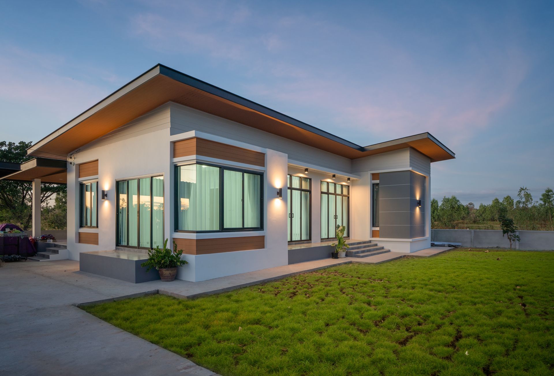 Modern, single-story house with a flat roof; exterior, dusk. Gray, white, brown accents, green lawn.