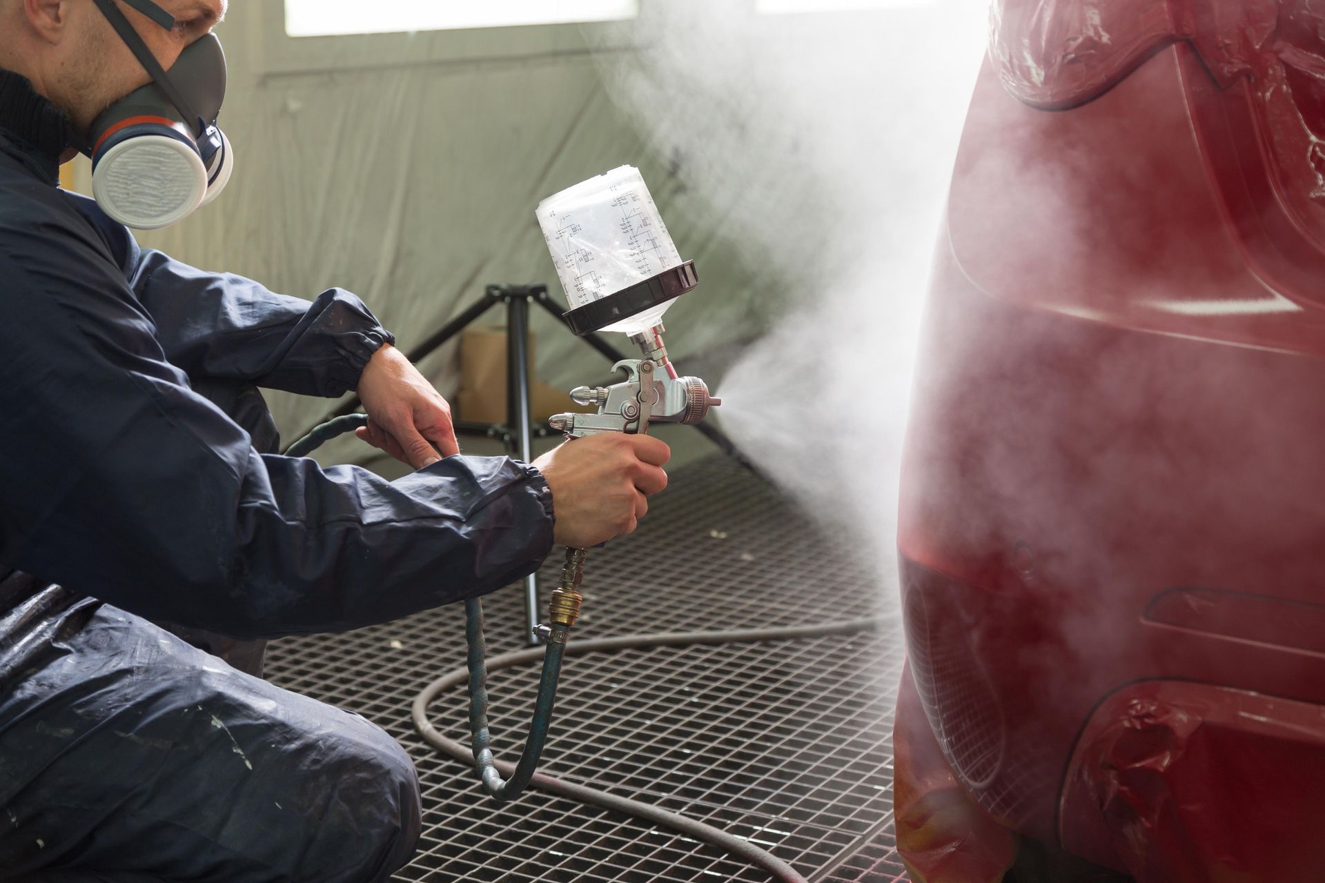 Person in respirator spray painting a red car panel in a paint booth.
