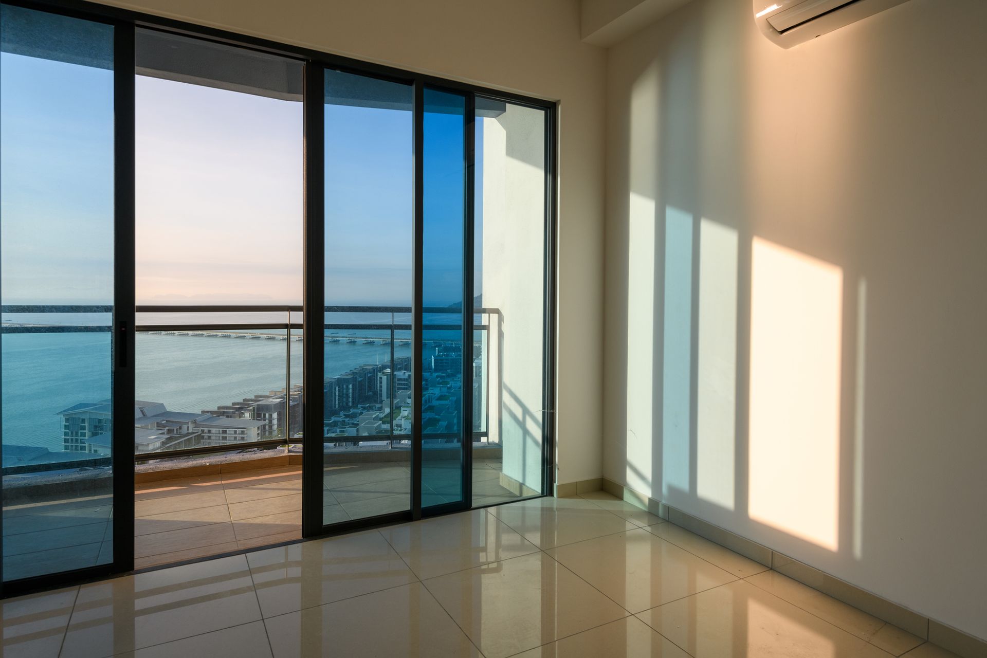 An empty room with glass doors to a balcony overlooking a city and ocean, sunlight on a tile floor.