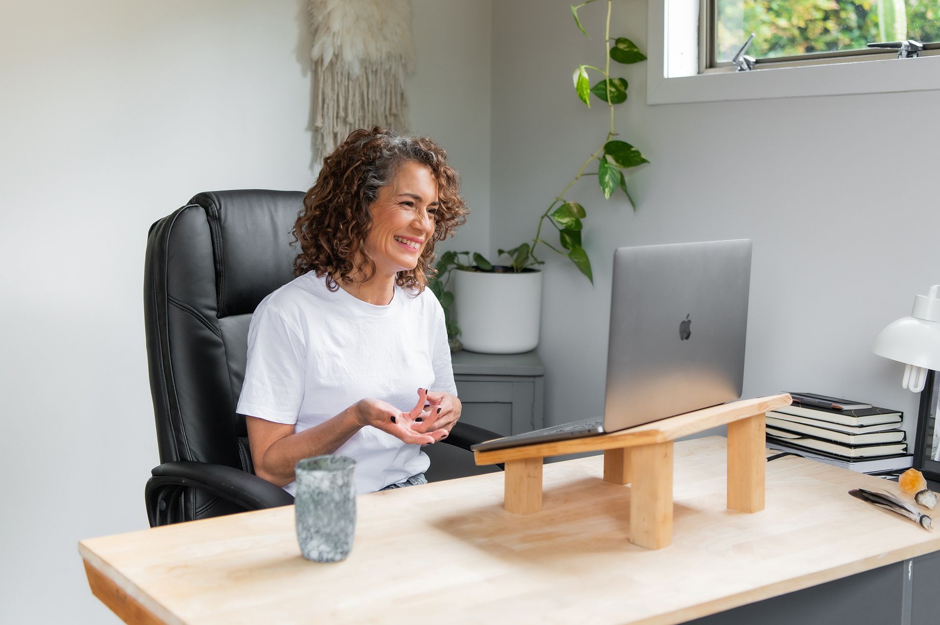 Tracy Manu with curly hair smiles while on a video call using a laptop on a wooden stand at a desk in a home office.