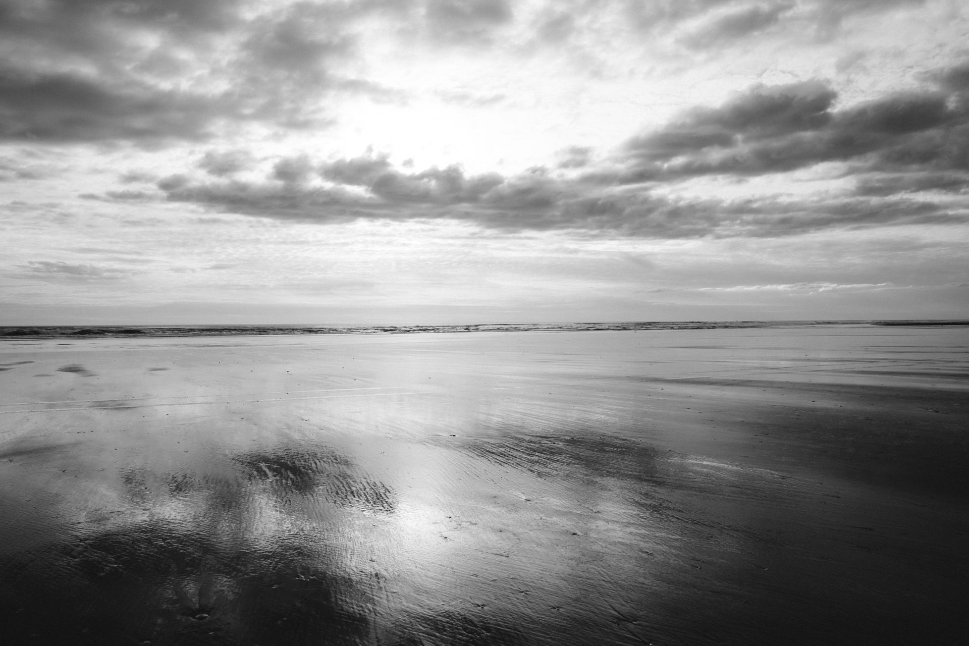 Black and white photo of a beach at low tide under a cloudy sky; wet sand reflects the clouds.