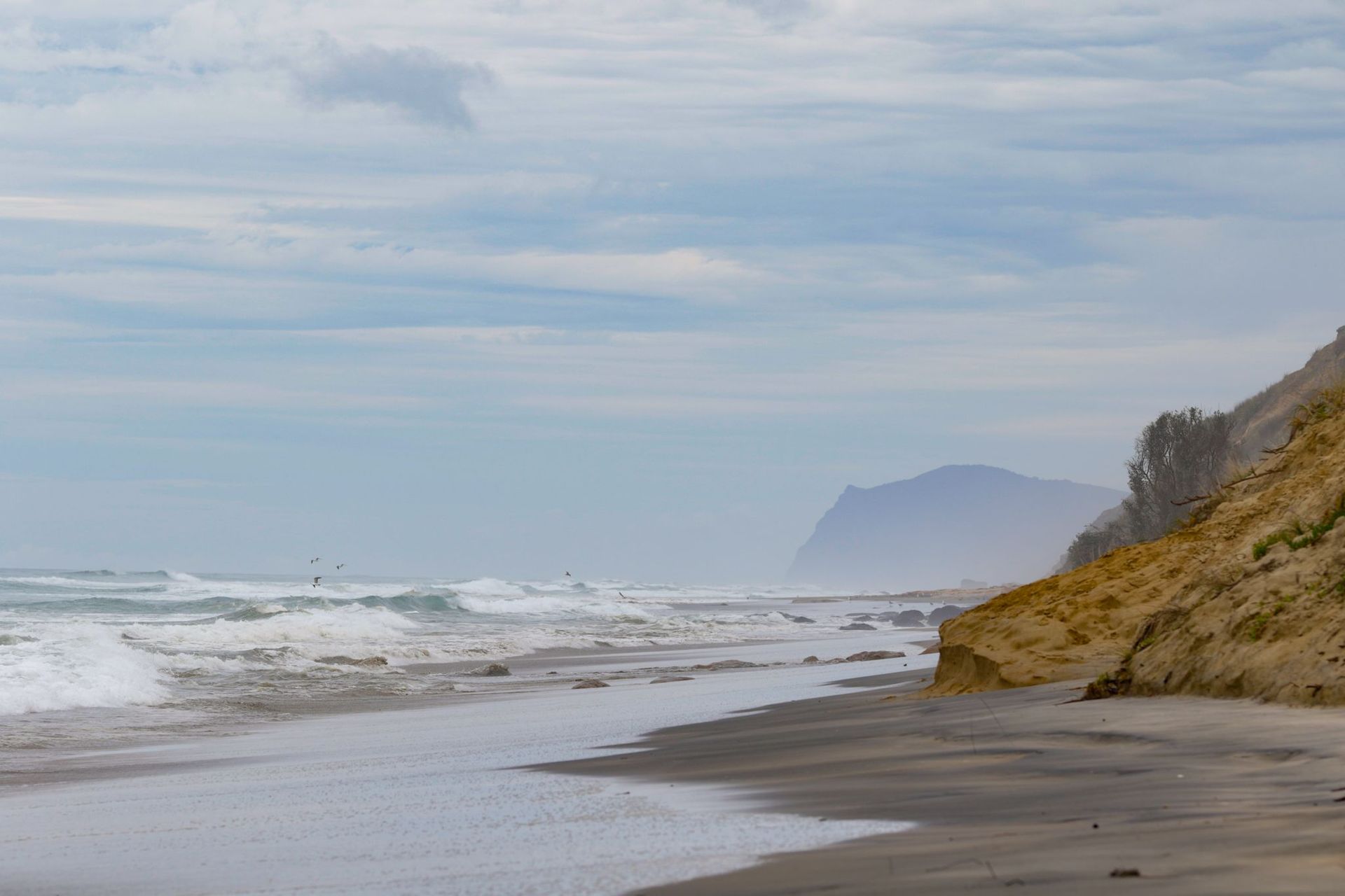 A cloudy beach scene with waves crashing onto the shore. A sandy cliff borders the right side with a mountain in the distant background.