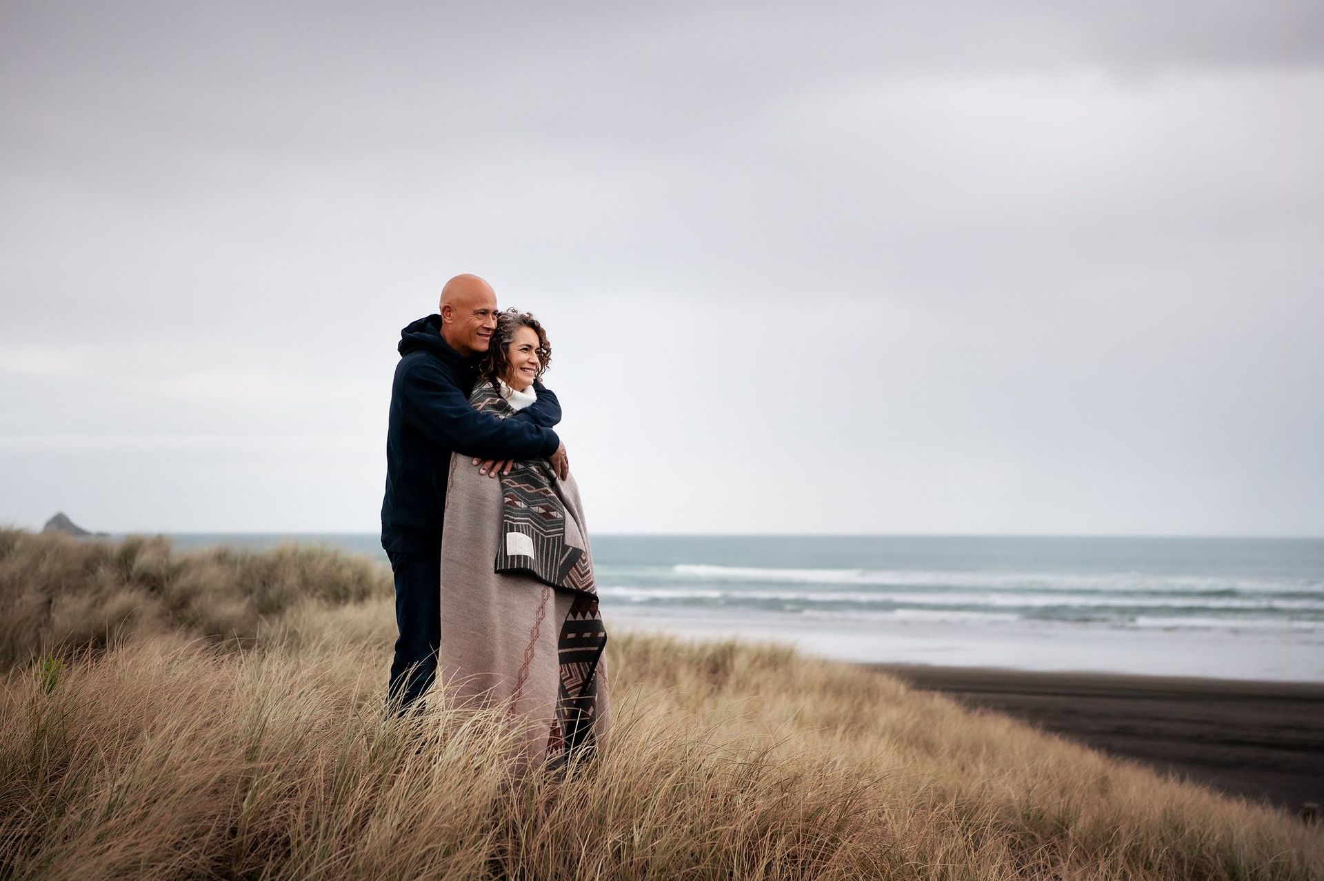 Tracy Manu and her husband embracing on a dune overlooking the ocean, wrapped in a blanket, against a cloudy sky.