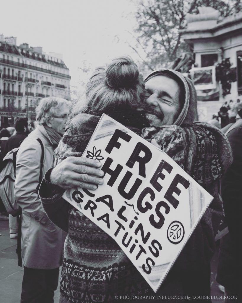 Person giving free hugs in a crowd, holding sign in English and French. Smiling faces, warm embrace.