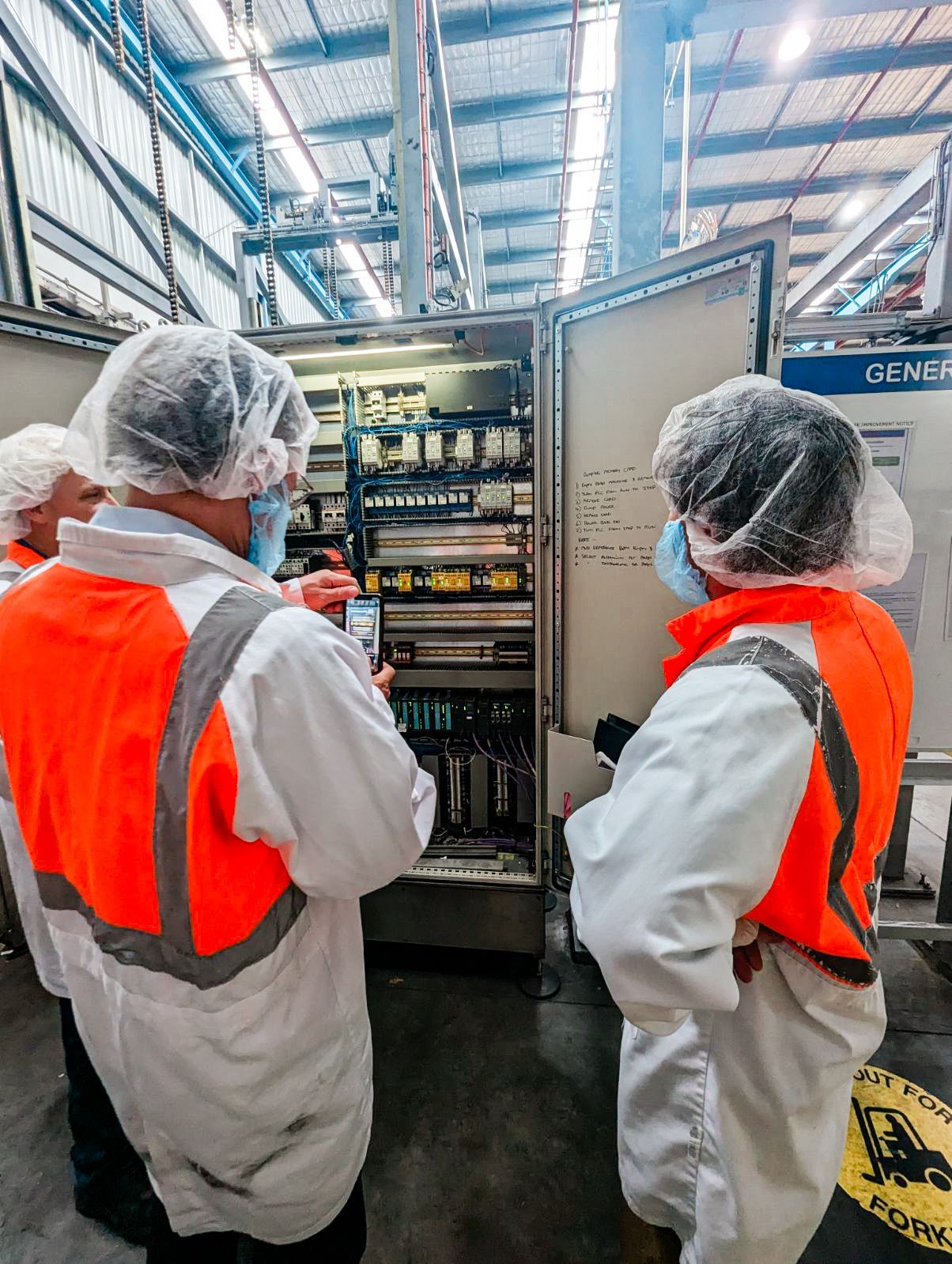 Two workers in protective gear examine machinery in a factory setting.