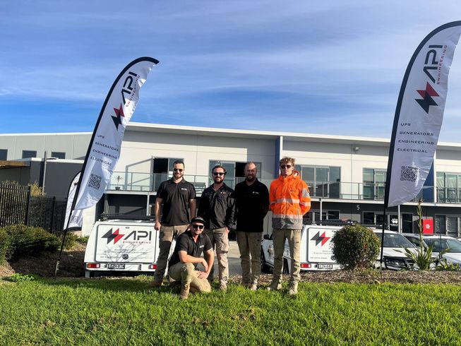 A group of five men pose in front of a building with flags and two trailers, all with the API logo.