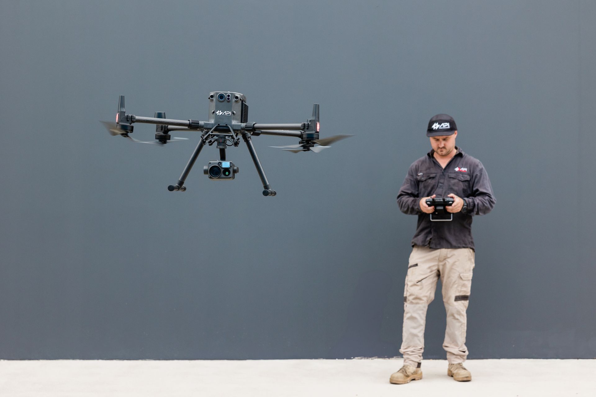 Man operating a drone against a gray wall. The drone is black, and the man wears a hat and work clothes. — API Engineering in Charmhaven, NSW
