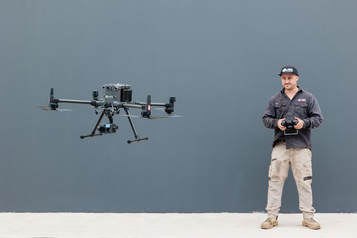 Man operating a drone in front of a gray wall; he's wearing a cap, work shirt, and cargo pants. The drone is black.