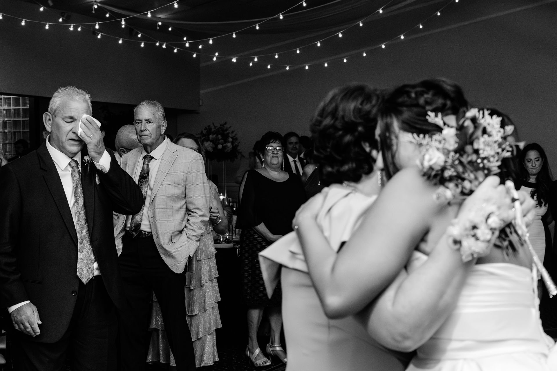 Wedding reception: man wiping tears, embracing bride, others watch. Black and white.
