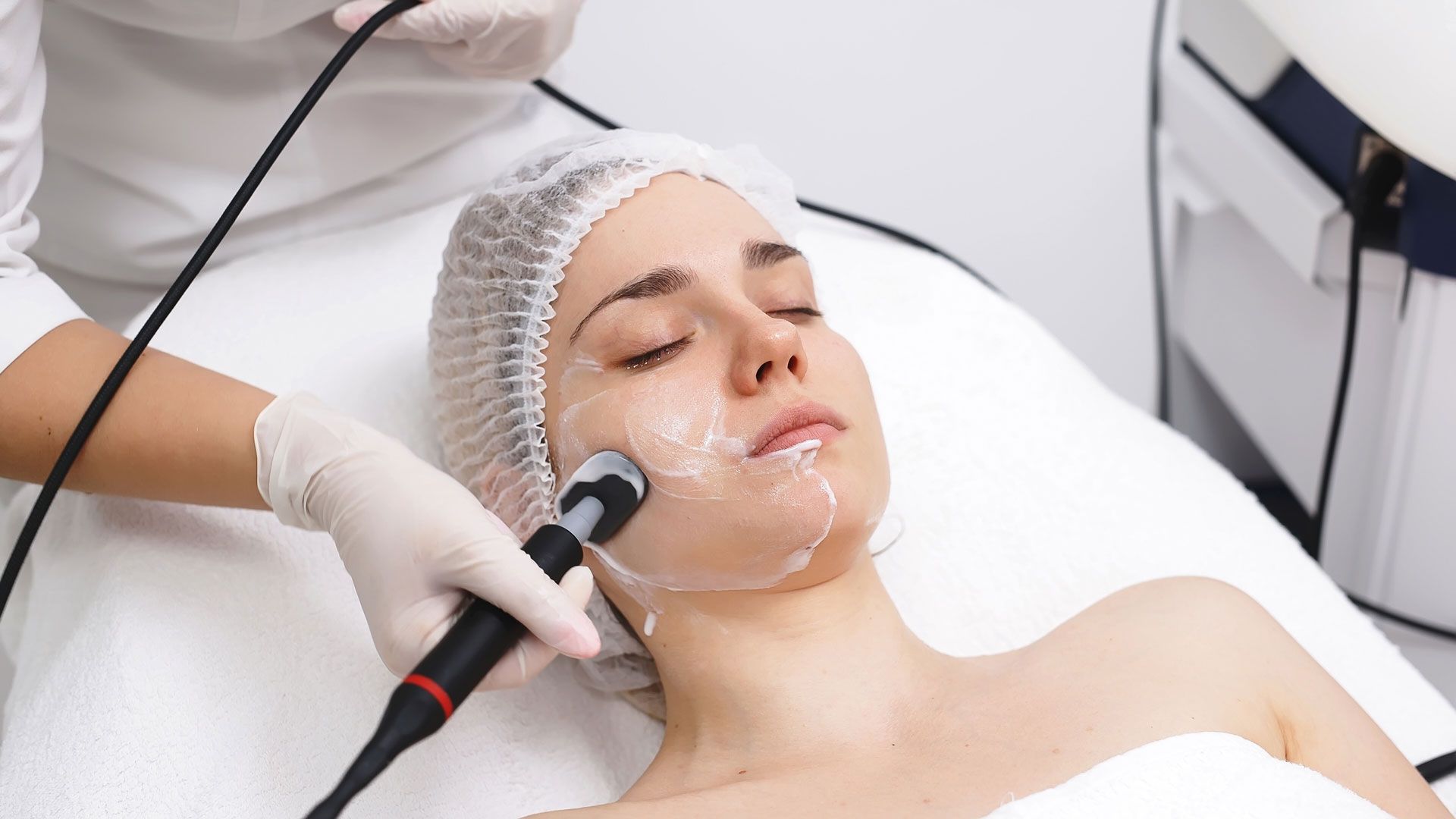 Facial treatment with a gloved practitioner using a handheld device on a woman’s face in a spa clinic