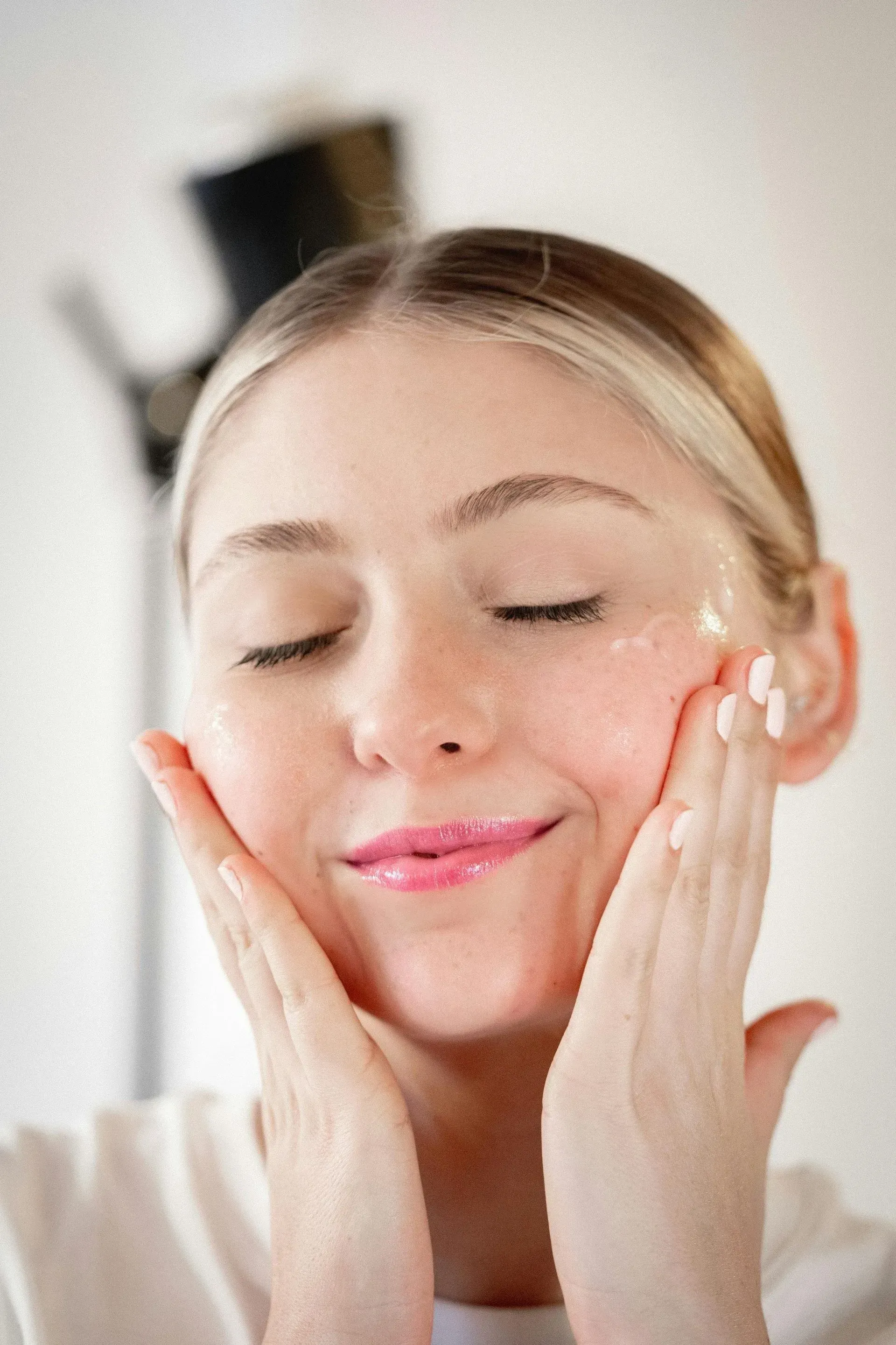 Woman gently applying facial moisturizer with both hands, eyes closed, in a bright bathroom