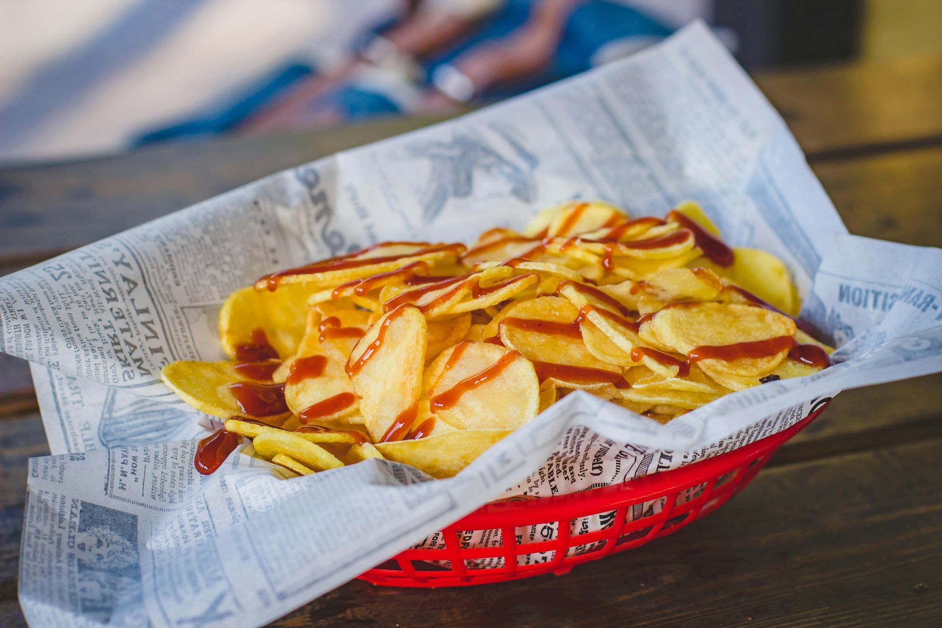 A basket of potato chips with ketchup on a newspaper on a table.