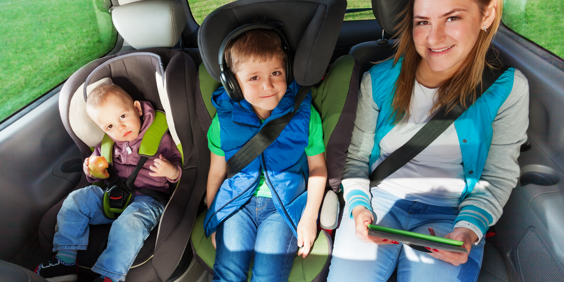 A woman is sitting in the back seat of a car with two children in car seats.