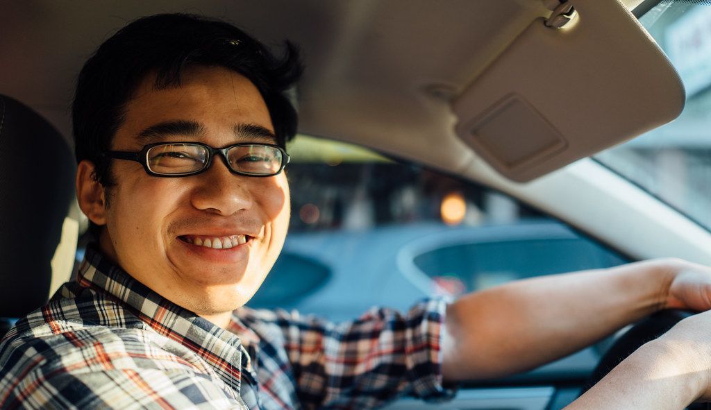 A man wearing glasses is driving a car and smiling.