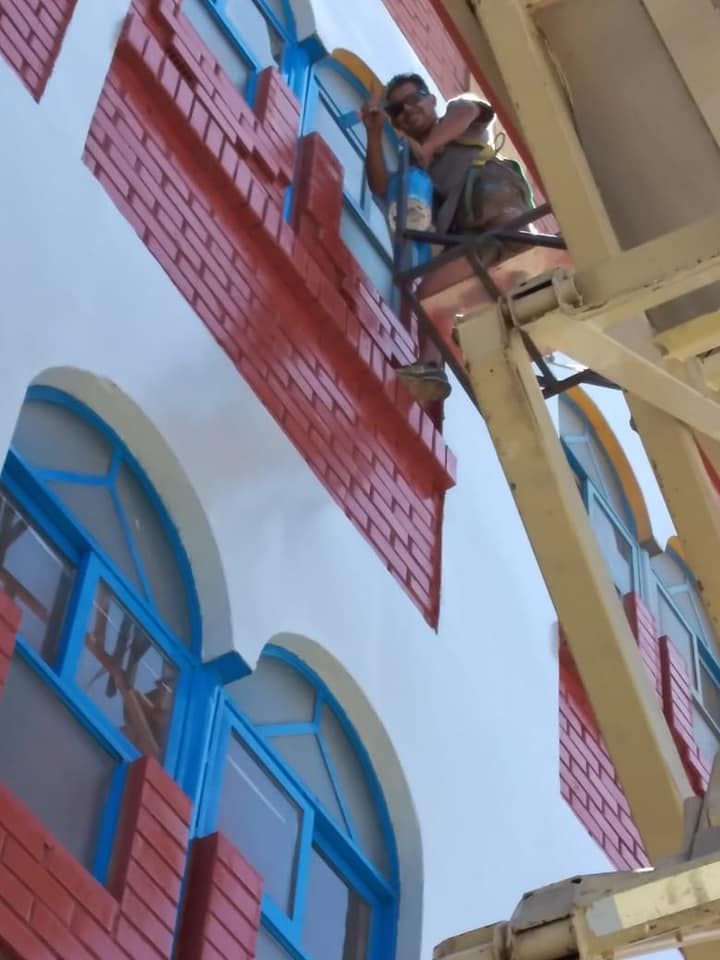 Persona pintando el exterior de un edificio con detalles de ladrillo y ventanas con marco azul.