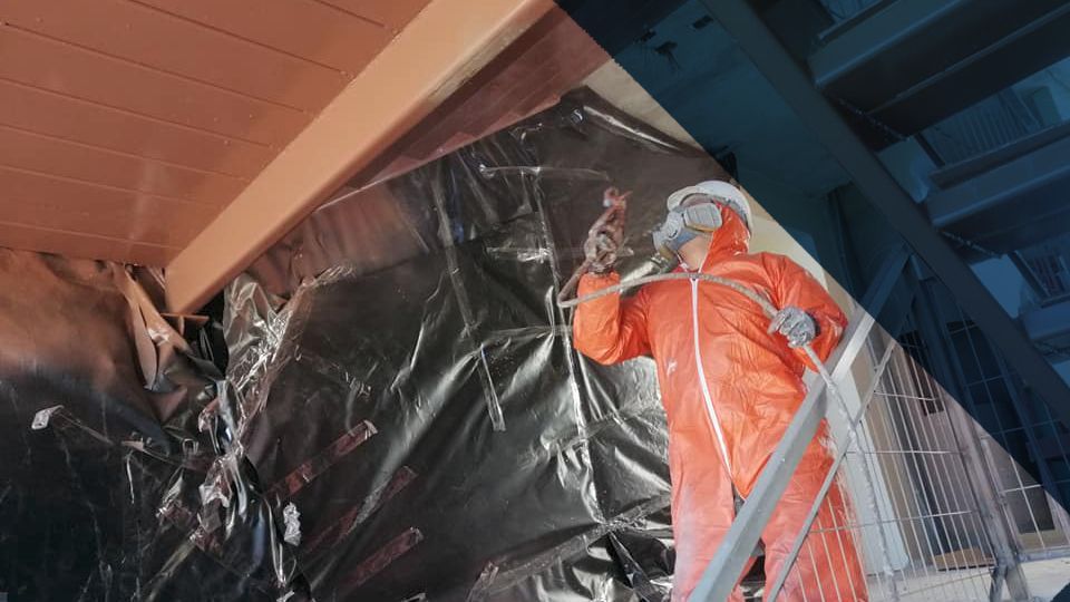 Trabajador con traje protector naranja pulveriza pintura sobre una viga cubierta de plástico negro.