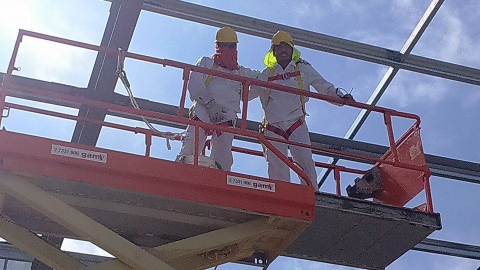 Dos trabajadores con equipo de seguridad en un ascensor naranja, instalando vigas metálicas bajo un cielo azul.