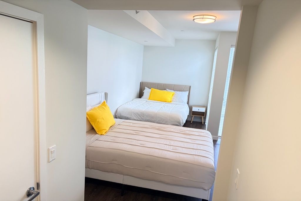 Small bedroom with two beds, white walls, and yellow pillows, viewed from the doorway