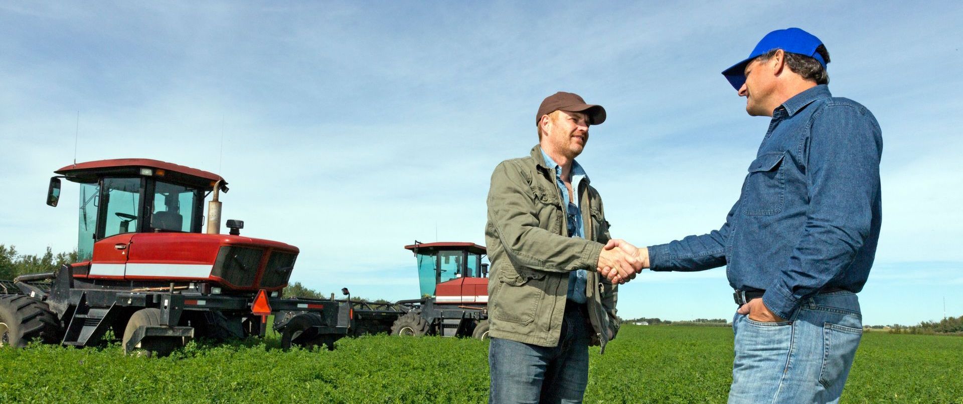 Two men are shaking hands in a field with tractors in the background.